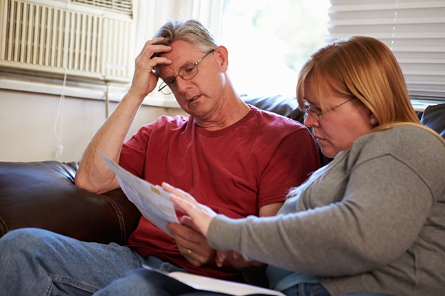 Worried Couple Sitting On Sofa Looking At Bills Worried Couple Sitting On Sofa Looking At Bills With Hand On Head